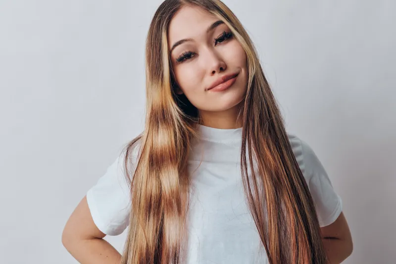 Portrait of confident young woman with long straight hair wearing white shirt standing with hands on hips posing against plain background indoors.