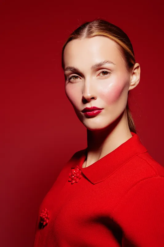 Portrait of confident woman with slicked hair wearing red sweater against red background. Studio lighting highlights her makeup with red lipstick and rosy cheeks. Concept of fashion and beauty.