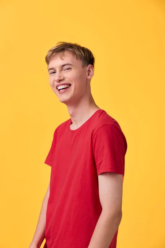 Smiling man in a red t shirt stands against a bright yellow backdrop, radiating casual energy, positivity, and youthfulness. The vibrant color contrast highlights a friendly, approachable vibe