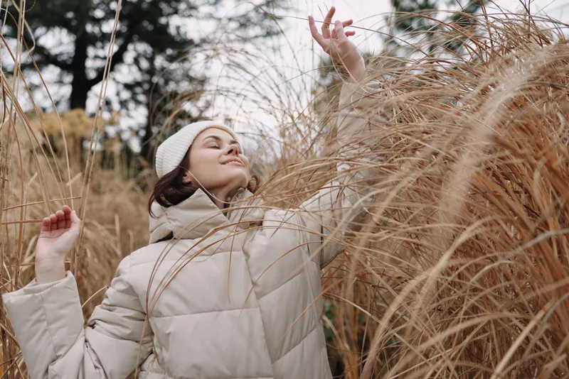 Woman wearing a light winter jacket and beanie stands among tall grasses in a breezy outdoor scene, smiling with arms raised, capturing joyful mood, wind swept hair and natural tones.