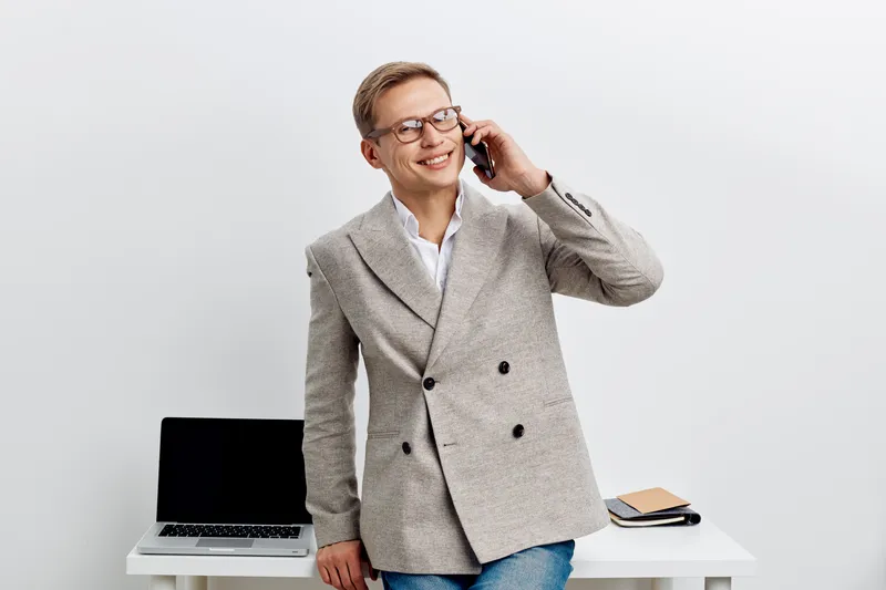 Smiling man wearing glasses, gray blazer, talking on mobile phone, standing at white desk with laptop and notebook, professional business indoor portrait, cheerful expression. People lifestyle concept