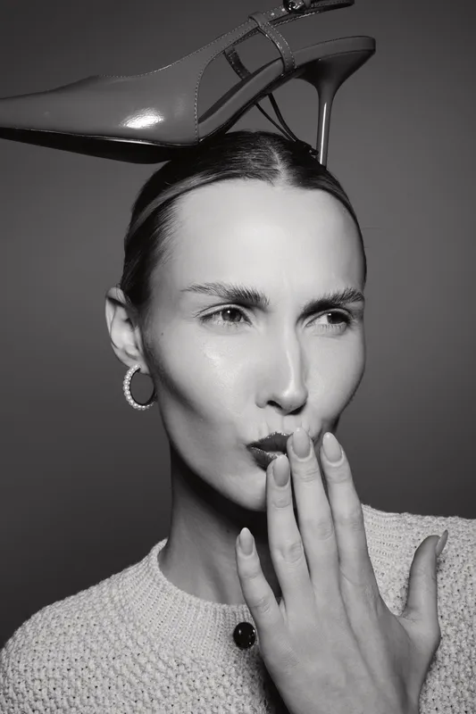 Caucasian woman with light skin and pulled back hair holds a high heel shoe on her head. She wears hoop earrings and a knit sweater in a black and white studio portrait.