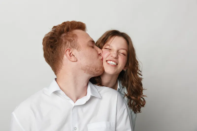 Young man kisses smiling woman on cheek in casual white shirts. Close-up portrait on light background. Concept of affection, love, happiness, and romantic relationship between couple.