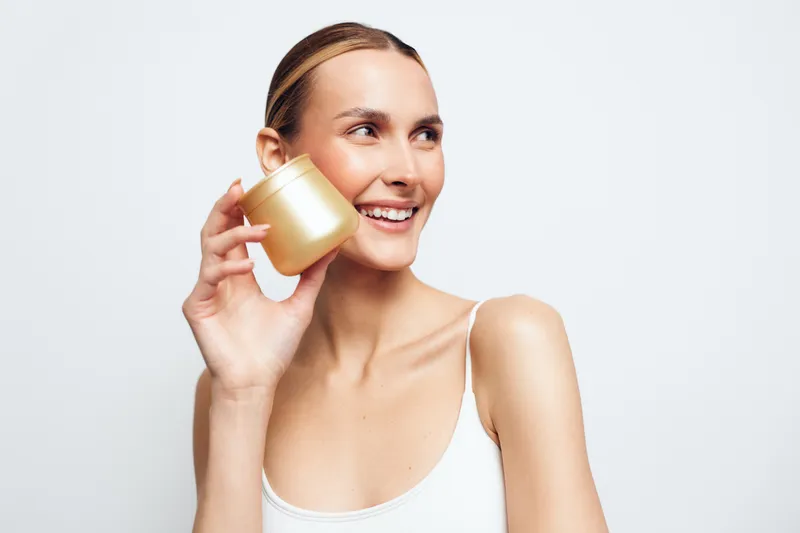 A smiling young woman with clear skin holds a cosmetic jar near her face on a white background. Concept for skincare, beauty routine, natural care, and wellness products.