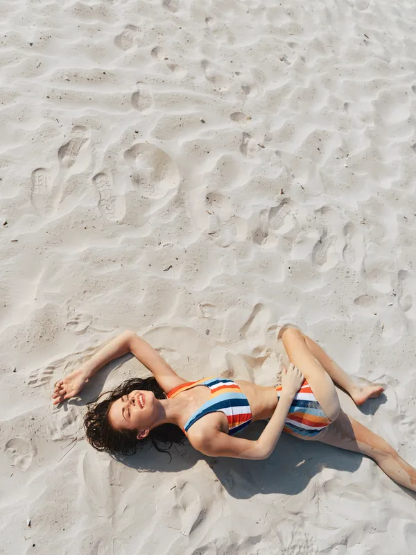 A young woman lies on bright sand in a striped swimsuit, relaxed with closed eyes. Sunlight highlights smooth skin, textured grains, and playful shadows, forming a carefree, summery moment of rest.