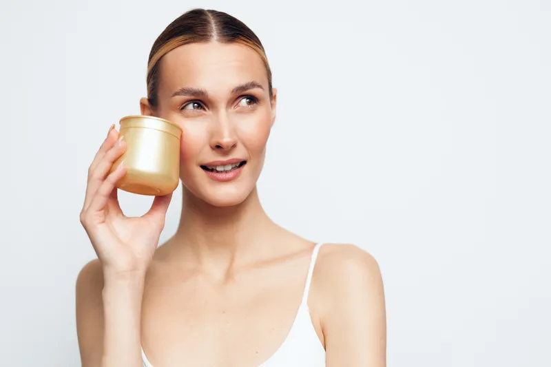 Young woman holding gold cosmetic jar near face, looking aside with slight smile, clean skin, white background, beauty product concept, skincare routine, natural look.