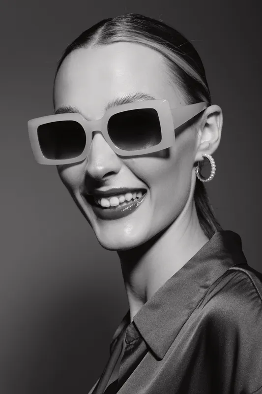 Smiling white woman with fair skin wearing large square sunglasses and hoop earrings poses in satin shirt on grey background. Concept for fashion, style, and accessories.