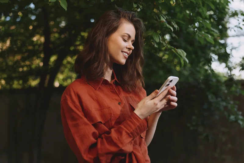 Woman lifestyle shot outdoors wearing a red shirt, smiling while using a smartphone, natural light, green trees in the background, relaxed candid moment in a park or garden setting