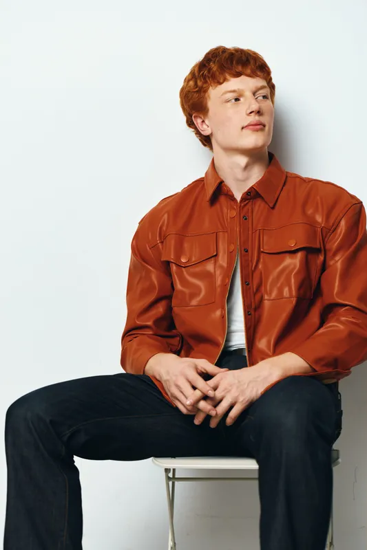 Young man with red hair sitting on white chair wearing brown jacket and dark jeans looking to side with thoughtful expression in studio against white background.