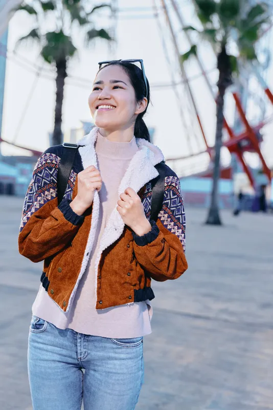 Outdoor fashion, smiling woman wearing a brown shearling jacket over a soft sweater on a sunny boardwalk, casual style, confident pose, palms in the background, travel vibe, fashion destination ready