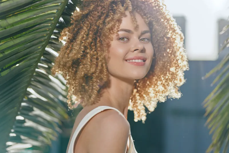 Smiling woman with curly hair stands outdoors between tropical palm leaves in soft sunlight, wearing a light summer dress and radiating a warm, happy vibe.