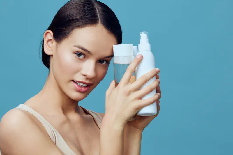 Young woman holding skincare products, showcasing beauty routine, smiling with confidence against a blue background, emphasizing healthy skin and hydration