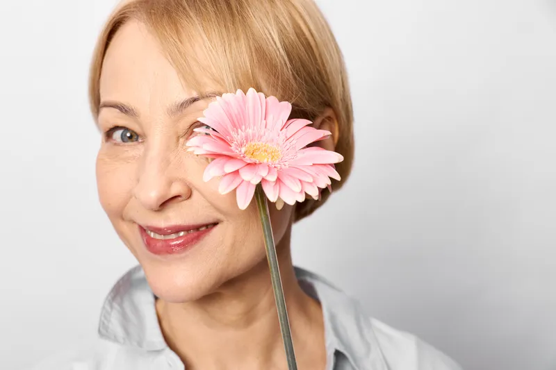 Cheerful senior woman closeup, smiling, holding a pink flower by her eye, studio setting with white background, soft light, gentle expression, natural beauty
