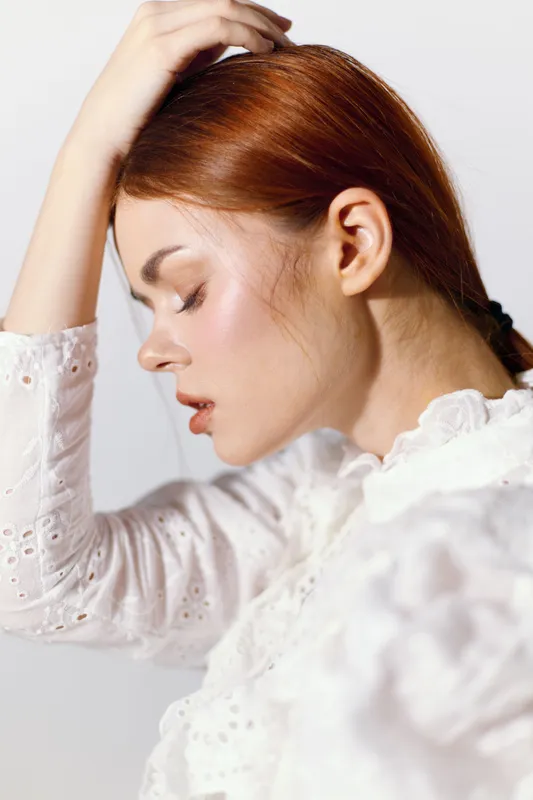Thoughtful young woman with red hair wearing a white blouse, expressing contemplation in a minimalistic light background setting