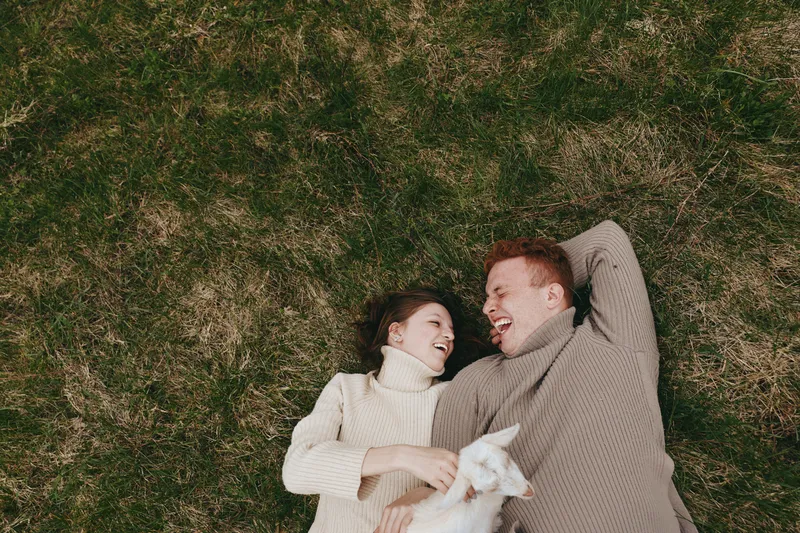 A couple lies on the grass outdoors, smiling and relaxed, with a small dog nearby. A warm, casual moment showcasing lifestyle, companionship, comfort, and simple joy in nature.