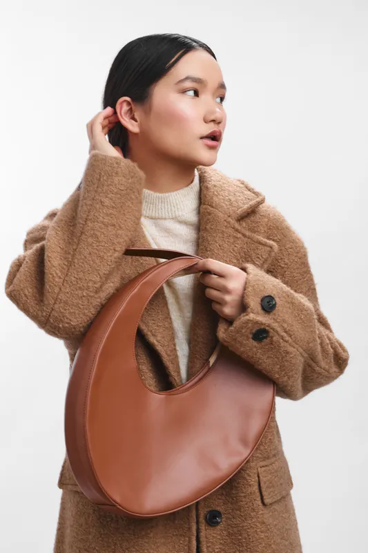 asian woman wearing a cozy brown coat and cream sweater stands with a curved leather bag, gazing to the side, conveying thoughtful style and calm confidence in a clean studio setting.