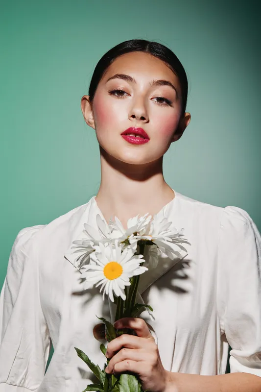 Young woman with vibrant red lipstick and soft glowing skin holds white daisies over her white dress. Clean background highlights beauty, fashion, and fresh creative style.