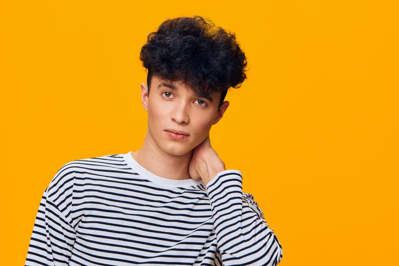 young man with curly hair in striped shirt poses thoughtfully against vibrant orange background, expressing calm and introspective mood in casual style