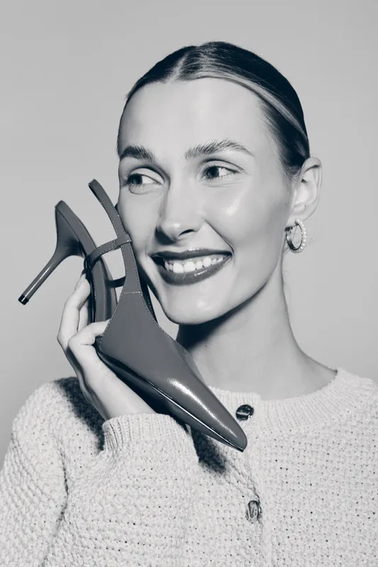 Young white woman with fair skin holds high heel shoe near face smiling, wearing knitted sweater and hoop earrings. Concept of fashion, beauty, and accessory.