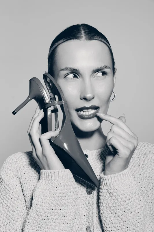 Young white woman with pale skin holds high heel shoe near face, looking sideways with thoughtful expression in black and white portrait. Concept of fashion and style.