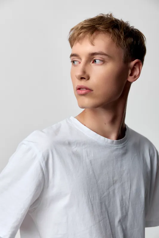 Man model in a profile pose looking away, wearing a white shirt under soft studio lighting, conveying calm, thoughtful mood with a minimal background, clean fashion vibe suitable for lifestyle,