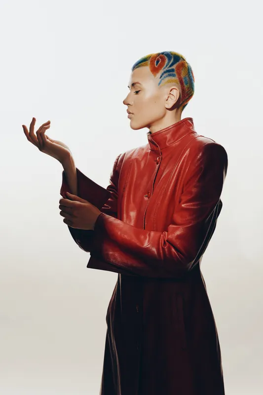 Woman with colorful short hair wearing a red leather jacket poses confidently against a bright neutral background in a modern fashion portrait.