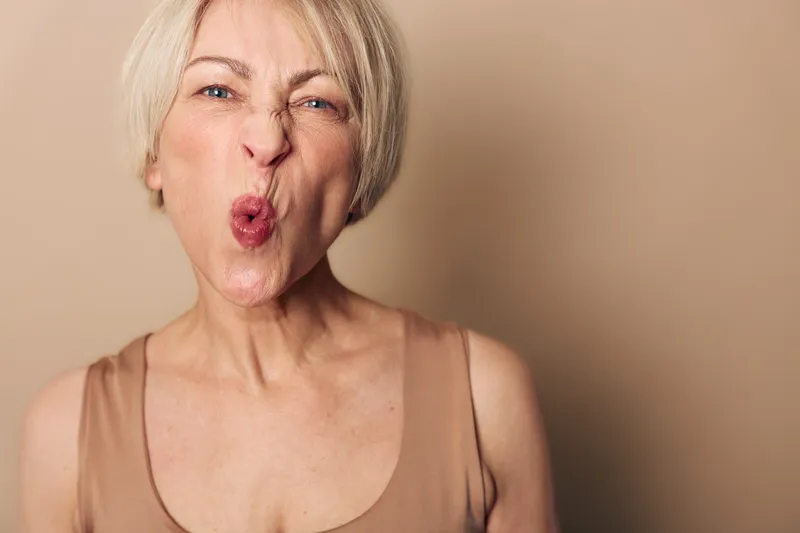 Portrait of mature woman with short blonde hair making a playful face, puckering lips in beige tank top against plain brown background. Concept for humor, expression, and natural aging.