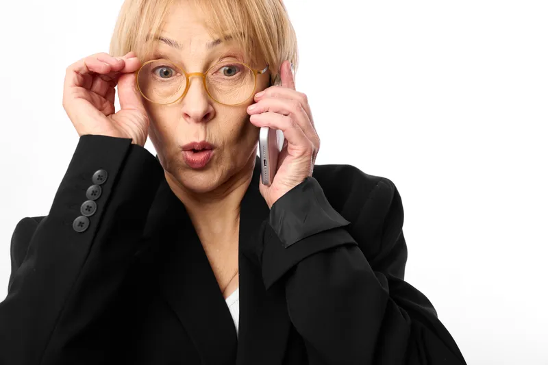 Elderly woman in business suit with glasses answers phone call, surprise expression, studio portrait on white background, formal blazer, professional communication