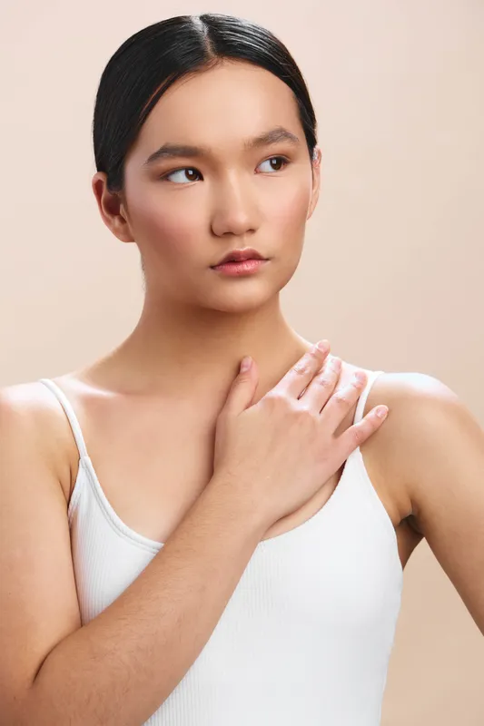 asian woman with calm expression stands against a soft beige backdrop, wearing a simple white tank top, conveying simplicity, poise, and gentle confidence in a studio portrait, suitable for
