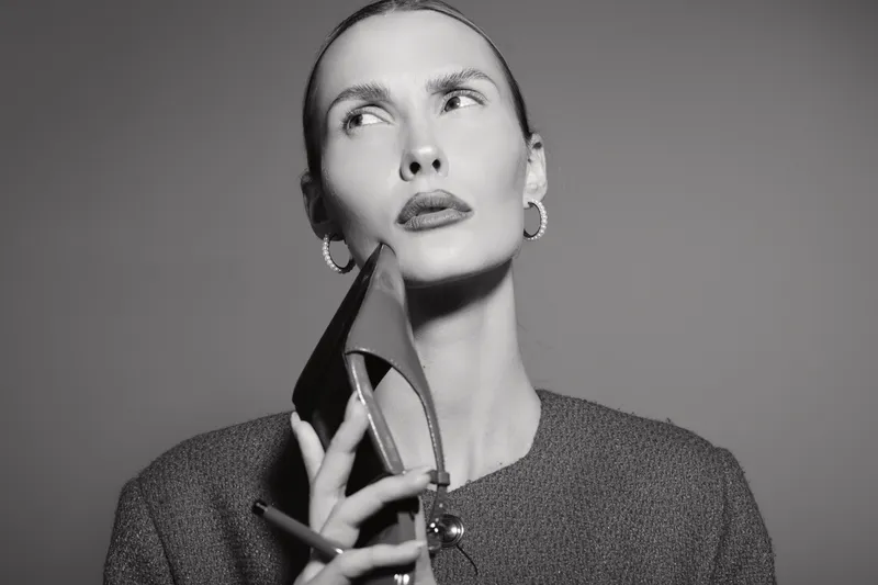Pensive white woman with pale skin holds high heel shoe near face in studio. Concept for fashion, style, and thoughtful mood in black and white portrait.