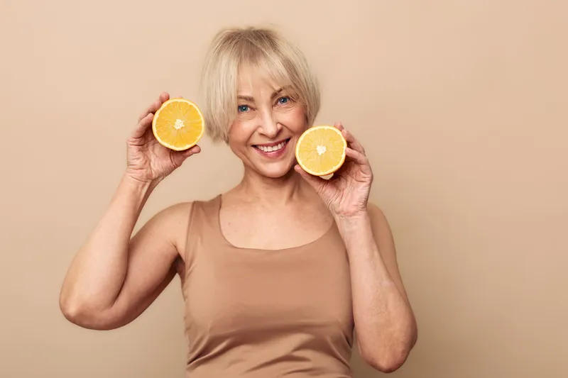 Smiling mature woman with blonde hair holds two orange halves near face on beige background. Concept of healthy lifestyle, vitamin c, nutrition, and natural beauty for older adults.