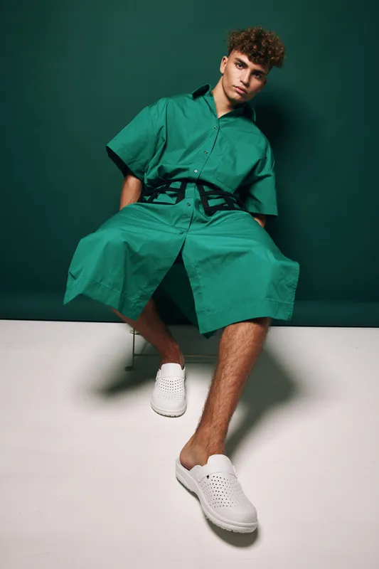 Young man with curly hair wearing oversized green outfit and white slip-on shoes sitting in chair against dark green background. Fashion, casual style and creative posing with urban vibe.