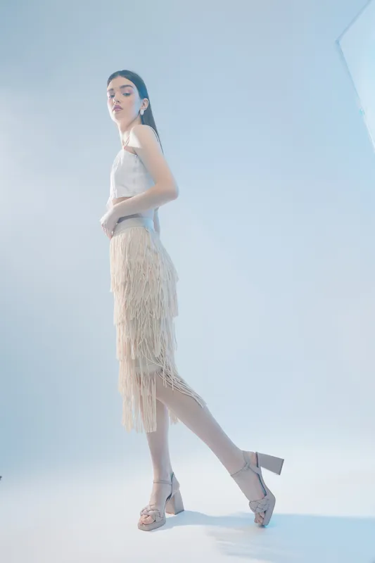 Young woman in a stylish beige fringe skirt and white top poses confidently in high heels on a light blue background. Concept of woman beauty fashion and elegance.