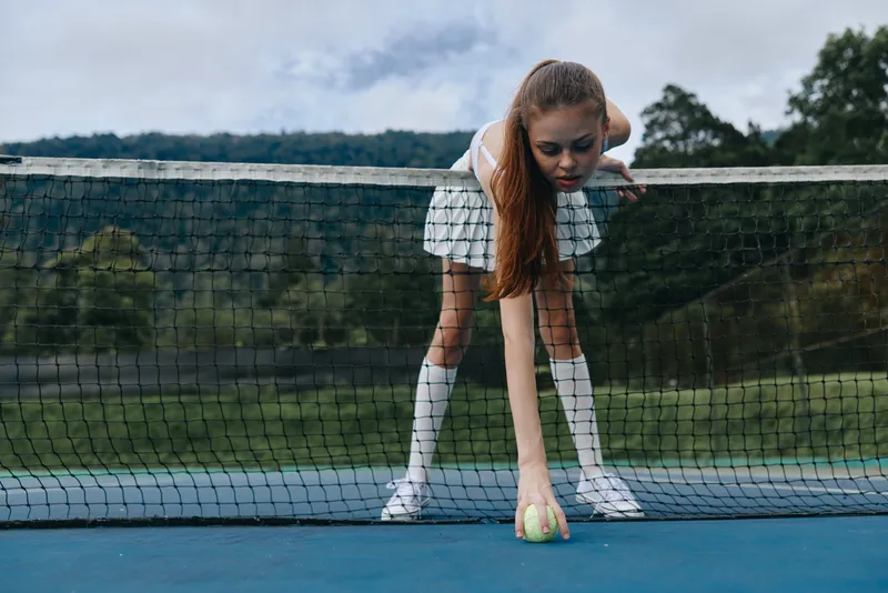 Young female tennis player in a skirt leaning down to pick up a tennis ball on the court, green nature background, showcasing concentration and focus