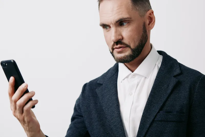 A professional businessman stands against an isolated white background, focused on a smartphone, projecting confidence, concentration, and calm determination, ready to lead with strategic insight and