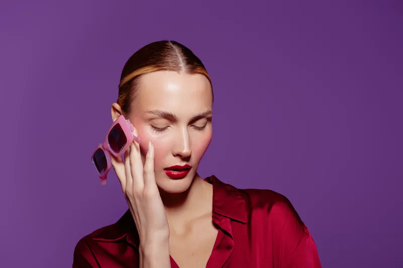 Young woman with slicked hair and red lipstick holds pink sunglasses near face with closed eyes on purple background. Concept of fashion, beauty, style, and elegance in studio.