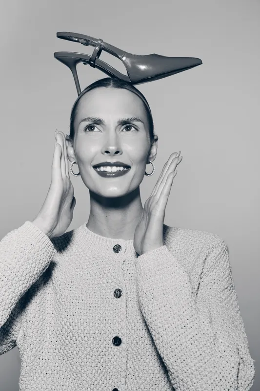 Smiling white woman with light skin wears a high heel shoe on her head, posing in knitted sweater and earrings. Concept of fashion, creativity, and playful style.