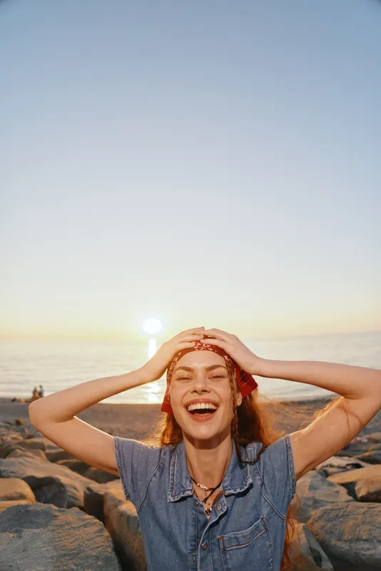 Lifestyle woman in modern boho-western denim street style laughing joyfully at sunset by the ocean, featuring warm film color tones and natural happiness.