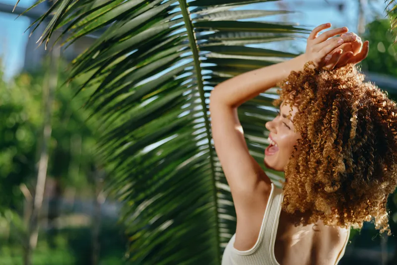 smiling woman with curly hair enjoying sunny tropical outdoors, wearing a white tank top, surrounded by green palm leaves, radiating happiness and warmth.