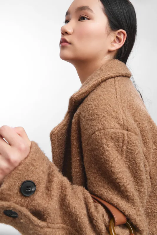 Asian woman in a warm brown coat gazes thoughtfully to the side, conveying calm confidence and poised professionalism in a minimalist studio portrait.