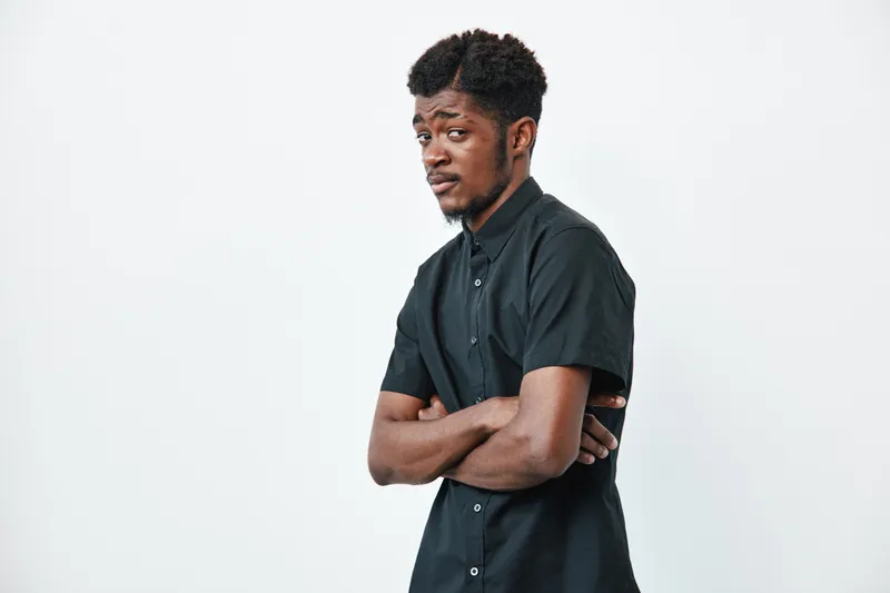 African American man with dark skin and curly hair wearing black shirt standing with arms crossed looking sideways on white background. Portrait, casual style and confident expression.