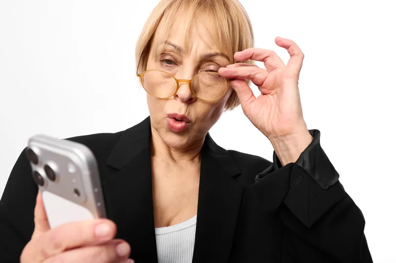 Senior business woman holds smartphone, adjusts glasses, surprised face, looking at screen, indoor studio portrait, communication technology concept, closeup.