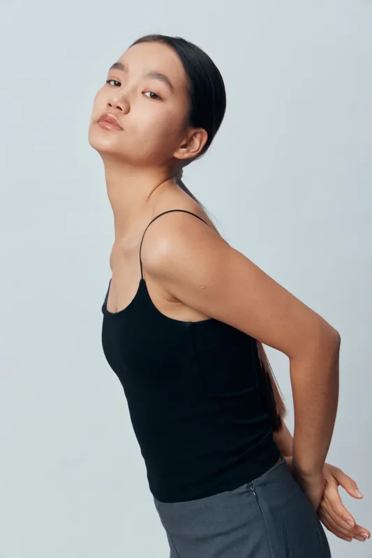 Young woman posing with head tilted and arms behind back wearing black tank top and gray pants on plain light background with serious facial expression and minimal makeup.