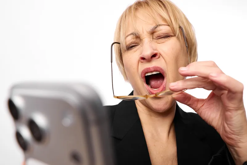 Tired businesswoman wearing a headset yawns as she talks into a smartphone, hand by her ear. Concept of call center burnout, exhaustion, and stress support.