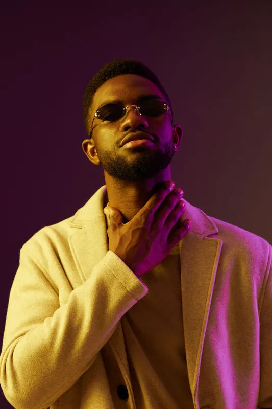 A stylish young man of African descent wearing sunglasses, a beige coat, and a confident expression against a purple backdrop, conveying elegance and modern fashion