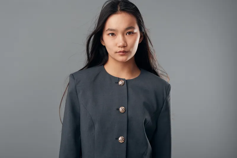 Young woman with long dark hair wearing a gray buttoned jacket standing against gray background looking at camera with confident expression. Studio portrait female model.