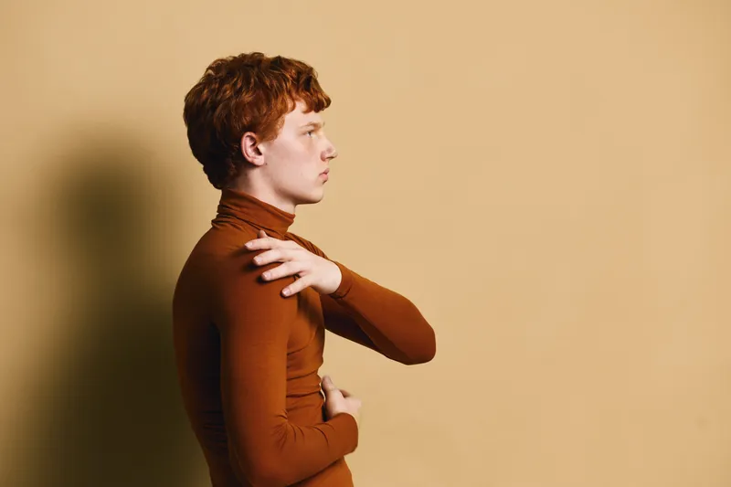 Profile of young man with red hair wearing brown turtleneck sweater standing against beige wall with serious expression and crossed arms.