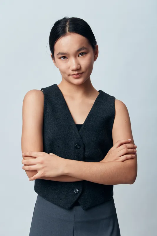 Young woman with crossed arms looking at camera with confident expression wearing sleeveless vest and dark skirt on plain light background. Studio portrait with natural makeup.