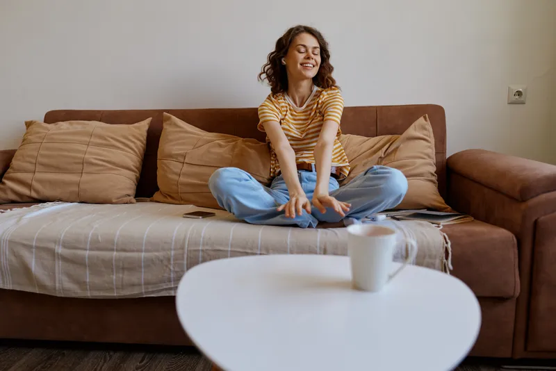A young woman sits cross legged on a brown sofa in a cozy living room, smiling softly as she reaches forward, enjoying a quiet morning.