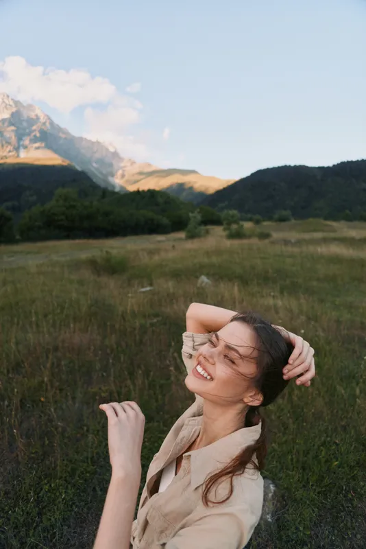 A woman outdoors in a sunlit field near mountains smiles with her eyes closed, casual beige shirt, wind in her hair. A lifestyle portrait of happiness and nature.